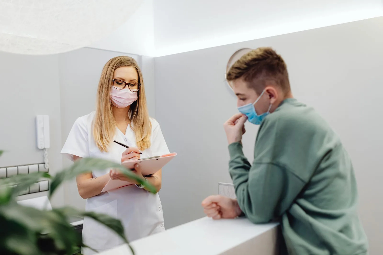 Un operatore sanitario consulta un paziente alla reception di una clinica, entrambi indossano mascherine.